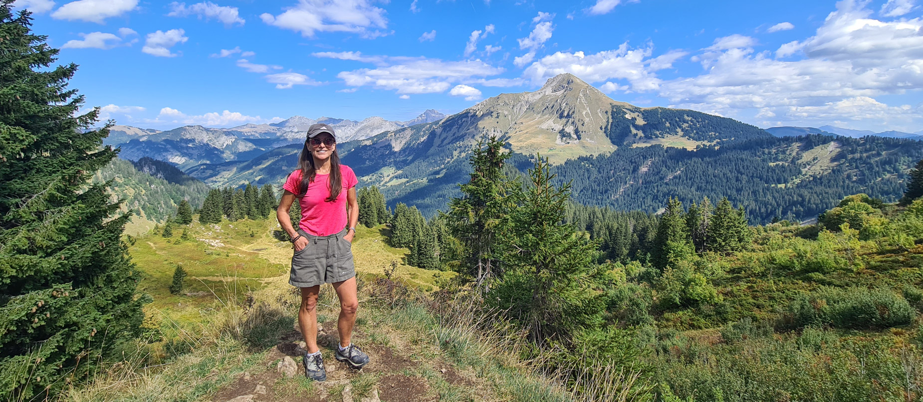 A professional solo female adventure traveler and photographer standing on a rugged alpine hiking trail in the Swiss Alps, looking out over dramatic mountain peaks and scenic valleys on a clear, bright summer day.