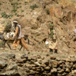 Local Afghan people walking a packed donkey along a narrow, rugged mountain trail cut into the rocky cliffs bordering the Panj River.