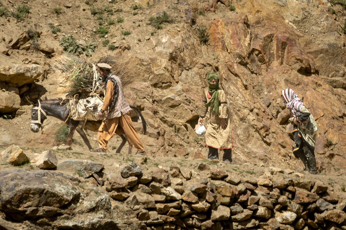Local Afghan people walking a packed donkey along a narrow, rugged mountain trail cut into the rocky cliffs bordering the Panj River.
