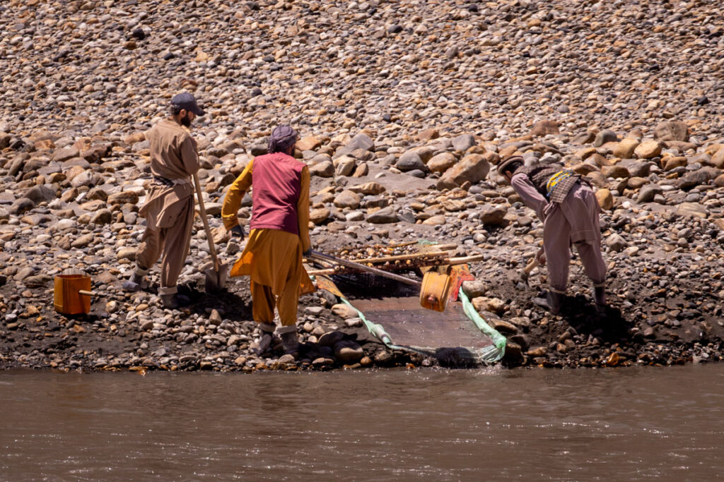 Men panning for gold using manual shovel methods and primitive mesh screens along the rocky banks of the Panj River border.